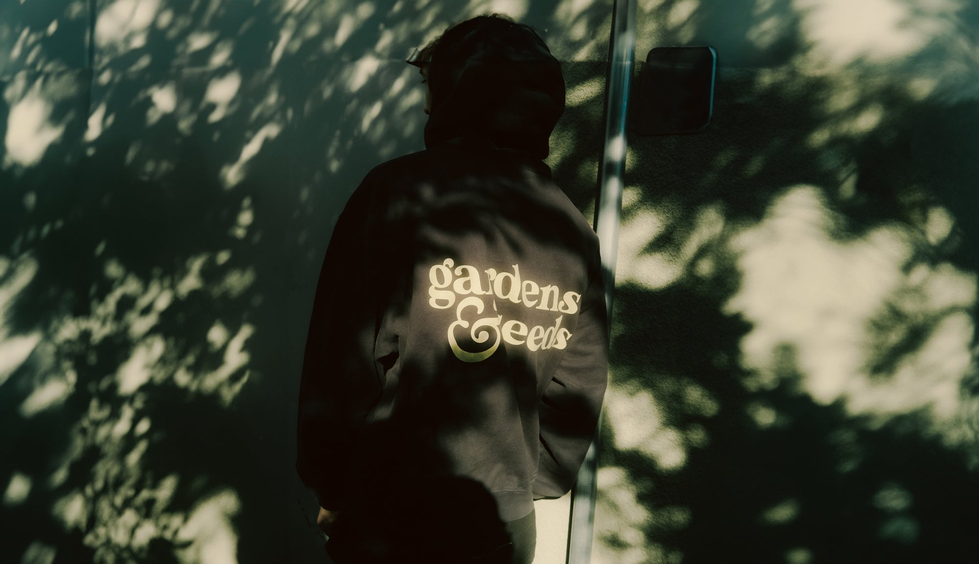 A man standing in the shade with a black Gardens & Seeds hoodie against a wall.