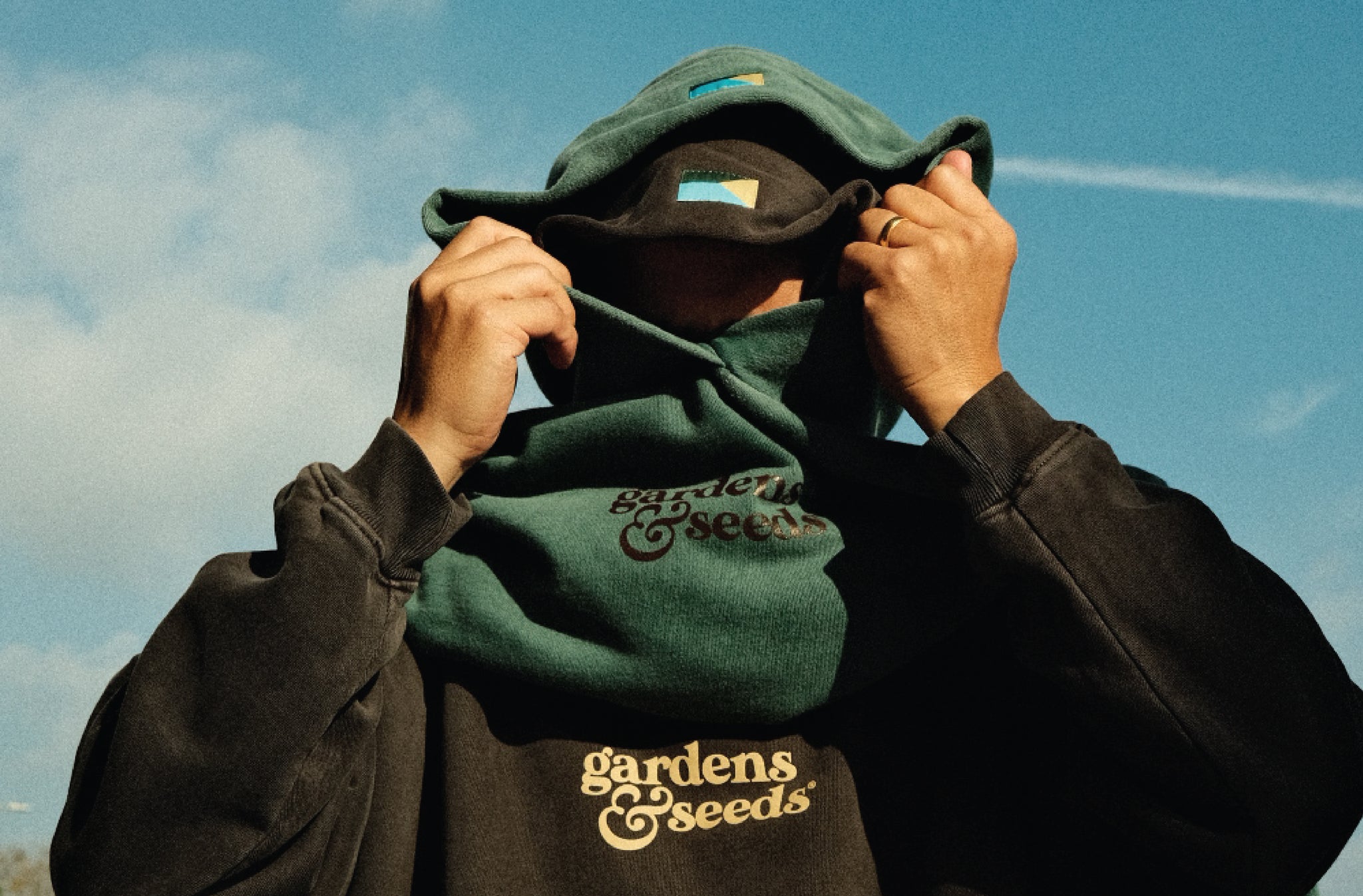 A man covering his face putting a Gardens & Seeds hoodie on top of another.